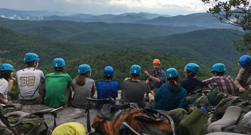 A group of people wearing helmets sit on an overlook, listening to a person speak. The blue ridge mountains are in the background. 